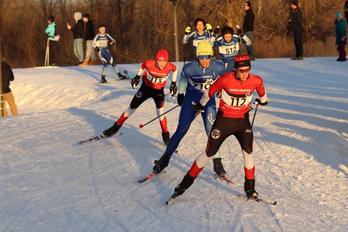 a small group of cross country skiers, including 2 in red and black uniforms, ski down a snow-covered hill with their shadows in the background