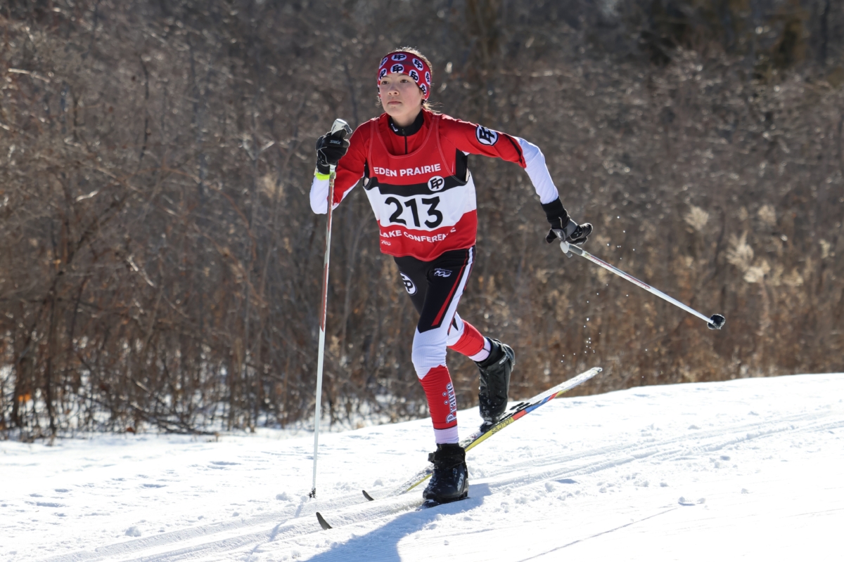 a young woman cross country skier in red and black uniform in mid-stride across snow