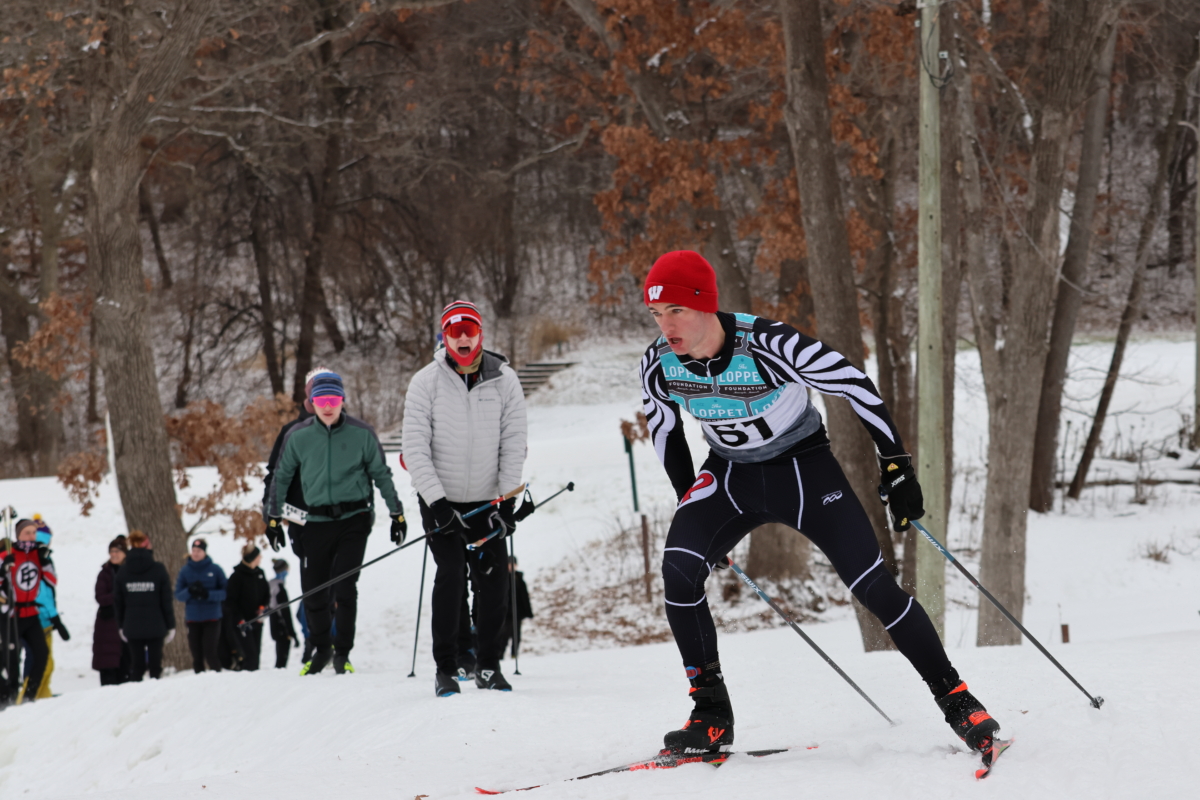 Young man cross country skier in red hat skis intently while spectators watch from below a slope.