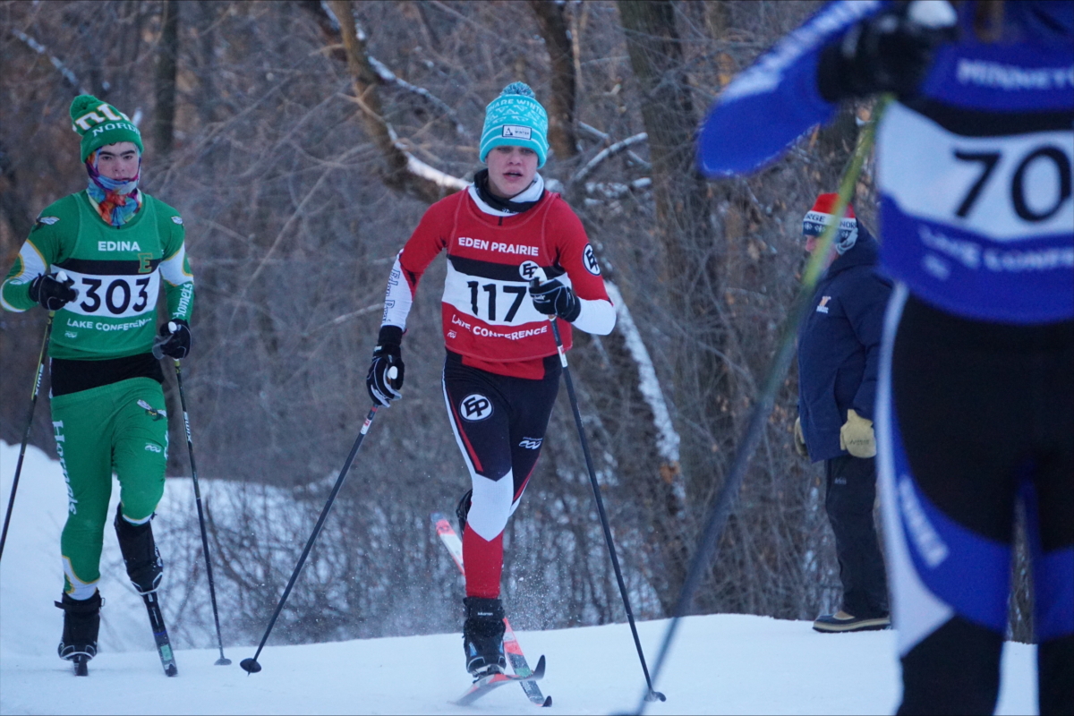 a young teenage boy in red and black uniform skis in front of another in a green and black uniform in a winter landscape