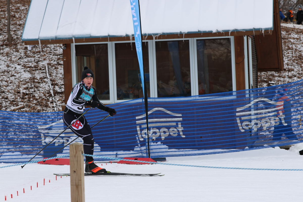 Young man cross country skier passes a building and blue loppet signs.