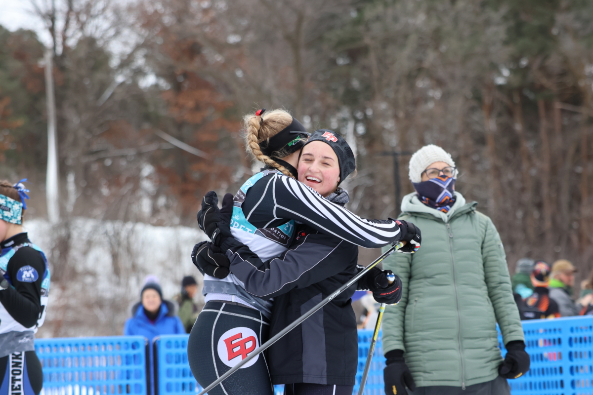 2 young women cross country skiers hug each other in front of blue net