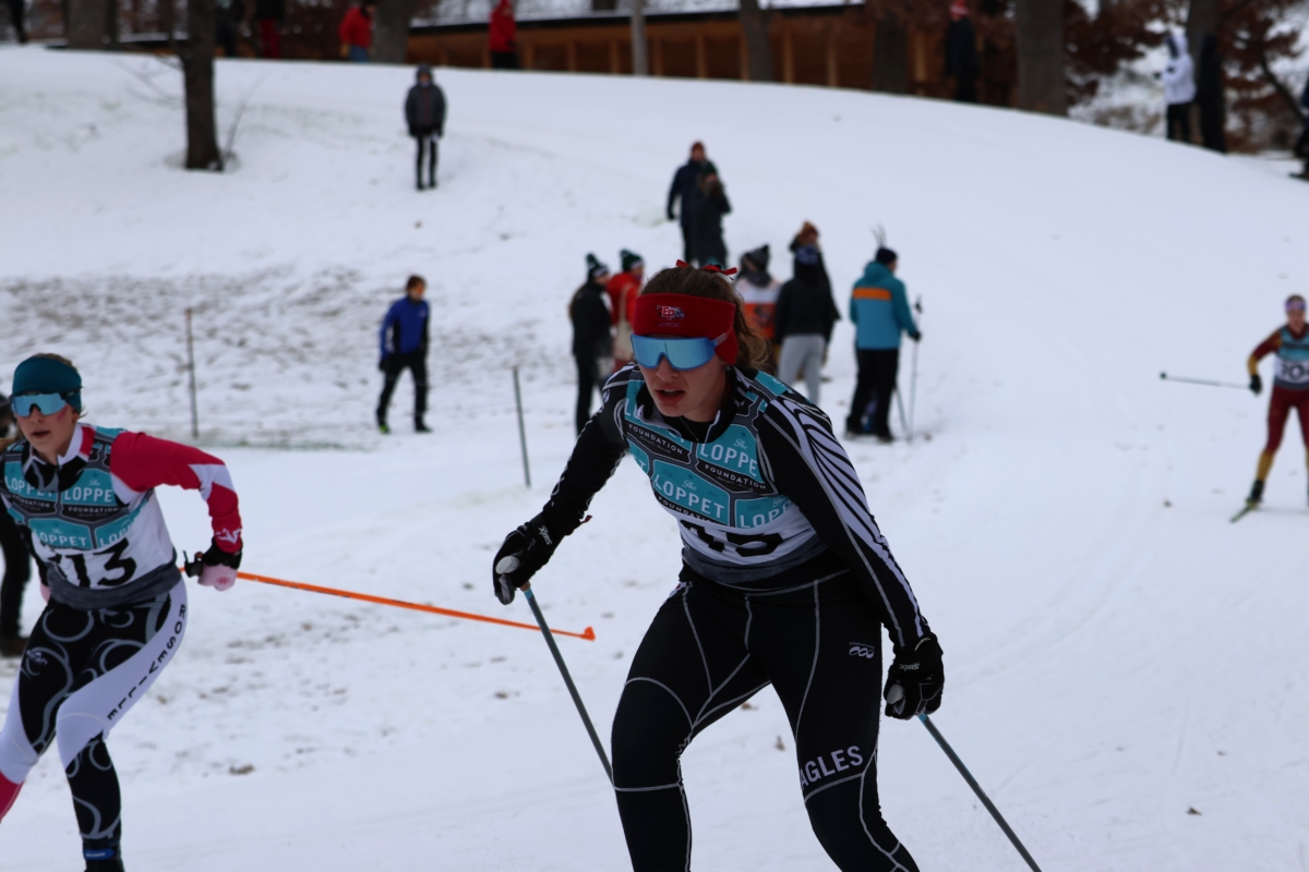 Young woman cross country skier in goggles with others visible on snow-covered hill behind her.