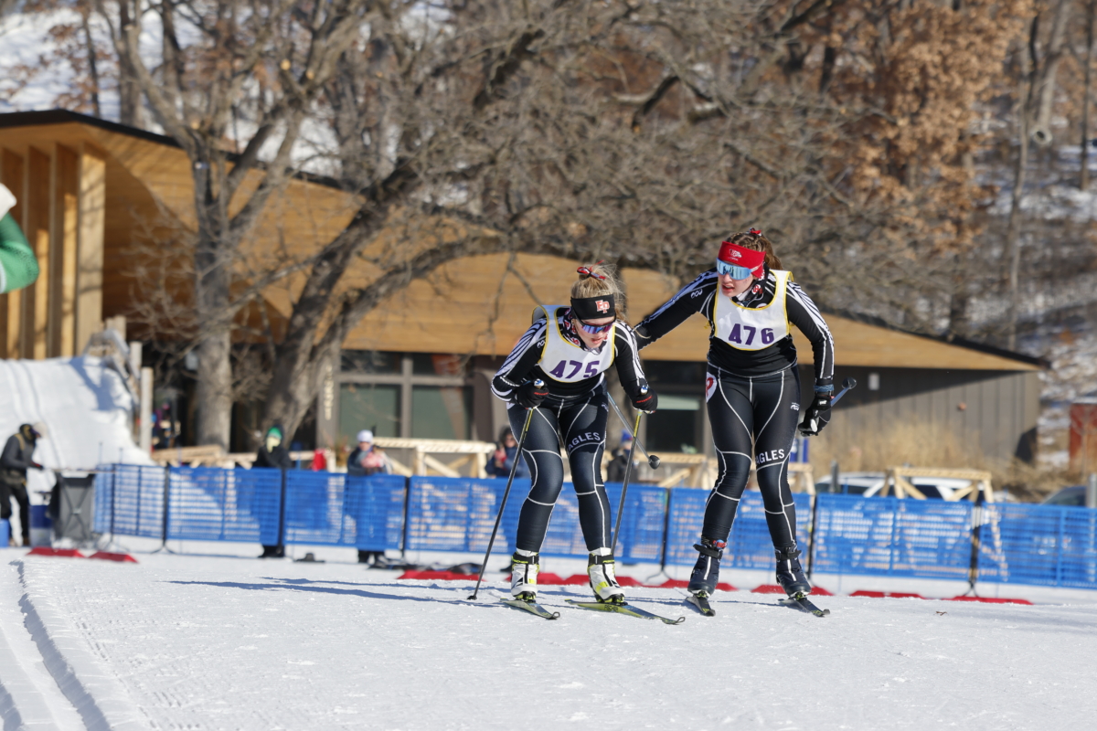 2 female skiers in black and white uniforms ski alongside each other on snowy slope in front of brown building