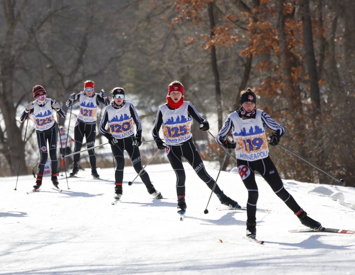 5 young female cross-country skiers in black and white uniforms ski with their poles in front of a treelike